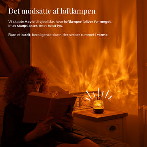 En kvinde læser ved siden af Havie, der spreder et varmt orange skær i et hyggeligt soveværelse.