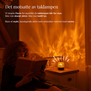 En kvinne leser ved siden av Havie, som sprer et varmt oransje lys i et koselig soverom.